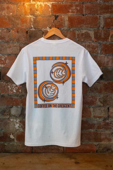 A white crew neck t-shirt with a geometric pattern in the form of latte art in coffee cups in orange and black colors, displayed on a hanger against a brick wall background.