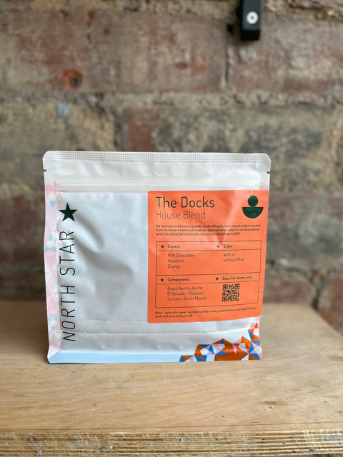 A packaged bag of 'North Star - The Docks' house blend coffee positioned against a brick wall backdrop on a wooden surface.